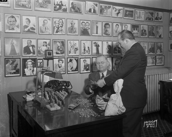 With a backdrop of celebrity portrait photographs and stacks of coins on his desk, Hugh Flannery, manager of the Orpheum Theatre, is receiving a $100 War Bond. The bonds were issued by the federal government to help finance World War II.