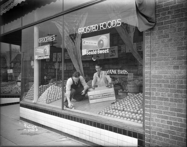 Frank Brothers grocery store, 609-613 University Avenue, with two workers stocking the window display with Seald Sweet oranges in neatly arranged pyramids.