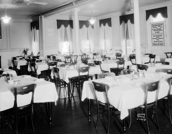 Dining room, Simon Hotel, 107-113 S. Butler Street, with tables set for dinner. A large plaque on the wall reads: "Eat More Cheese. Eat all You Want. No Extra Charge. We assist Wisconsin's Greatest Industry, Dairying. Using Wisconsin's products only." Owner A.M. Ophaug, proprietor.