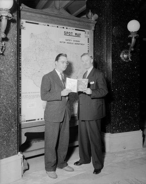 Secretary of State Fred R. Zimmerman and Robert "Bob" Brayton, Public Relations Director or the American Automobile Association. They are standing in front of a map in the Rotunda of the Wisconsin State Capitol that shows the Wisconsin traffic fatalities for the years 1939-1940.  Zimmerman is present for this photo opportunity because Wisconsin drivers licenses were then issued by the Secretary of State. The name recognition Zimmerman gained from having his signature on the licenses made him one of the most electable candidates in Wisconsin history.