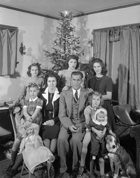 Group portrait of Denton family showing parents, five daughters, two dolls, a Christmas tree and a dog.