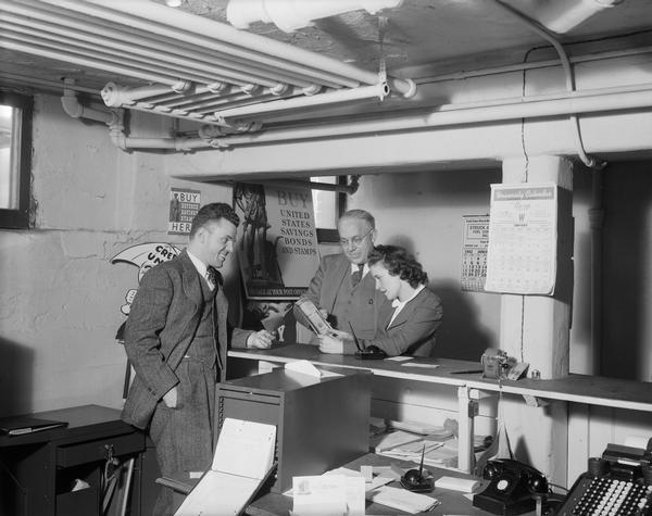 Man and woman consider buying Defense Savings Bonds and Stamps from a clerk at a basement CUNA Credit Union National Association office.