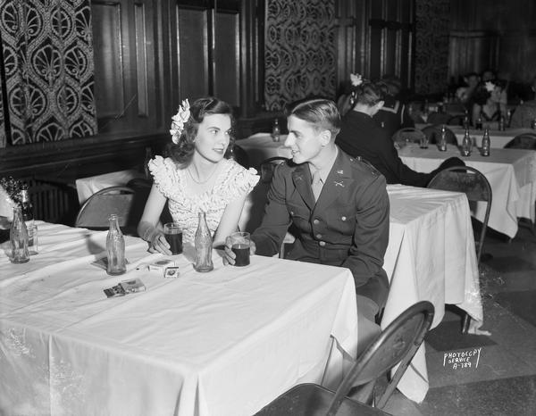 A formally dressed young woman and her beau in his U.S. Army uniform sit at a table and drink Coca-Cola at the "Moonlight Formal" in Tripp Commons at the Memorial Union on the University of Wisconsin-Madison campus.