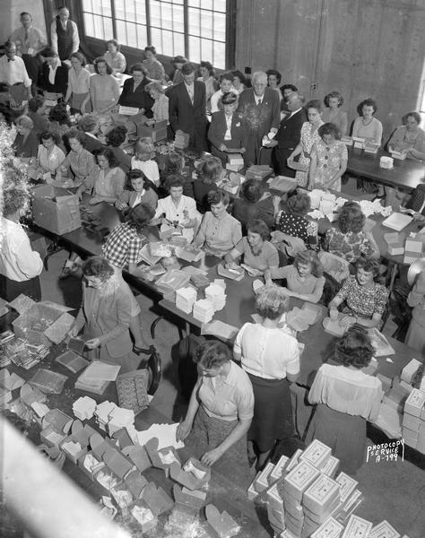 Elevated view of a large group of state employees are standing at long tables laden with items to be packed into Red Cross Christmas boxes for soldiers. The glassed in, lakeside basement in the rear of the State Office Building was used for the assembly line. Governor and Mrs. Goodland can be seen near the rear window viewing the work.