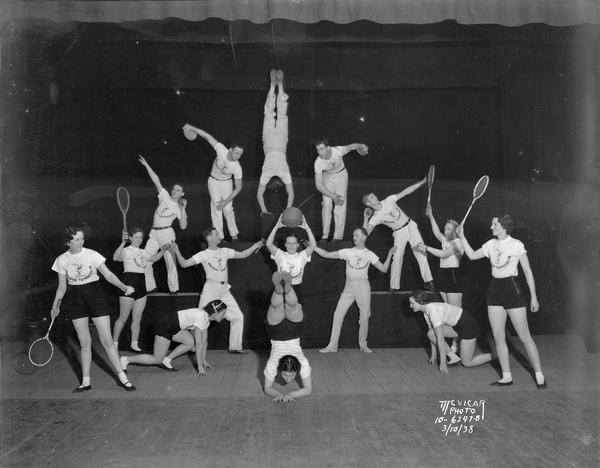 Acrobats in formation, on stage with 15 participants holding various pieces of athletic equipment.
