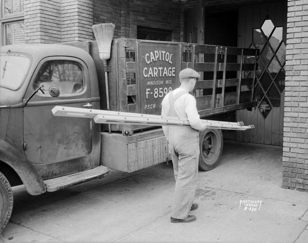 Man in work clothes carrying a folding ladder past a Capitol Cartage truck parked at Straus Printing Company.