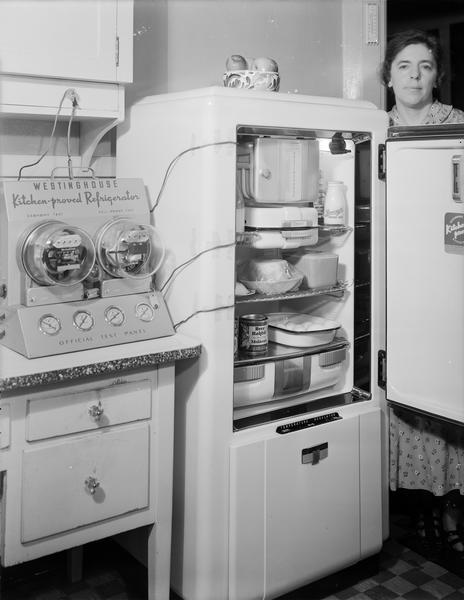 Testing a Westinghouse refrigerator (door open with food inside and woman standing behind the open door) with wires from an Official Test Panel attached to the refrigerator showing two dials for economy test and full power test, in house at 501 Welch Avenue.