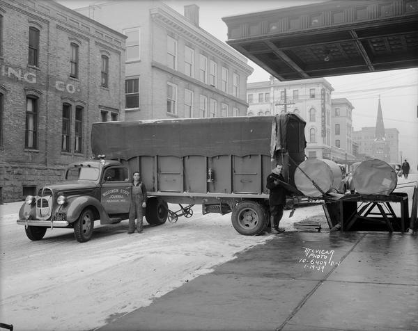 Men with tractor-truck unloading large rolls of paper at the Wisconsin State Journal, 115 South Carroll Street. Grace Episcopal Church, 110 W. Washington, the Park Hotel, 22 S. Carroll Street, the Lamb Building, 105-109 W. Main Street, and the Democrat Building, 114-124 S. Carroll Street can also be seen in this view.