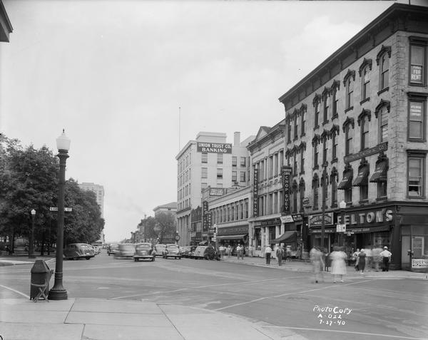 View of businesses located on West Main Street at S. Carroll Street on the Capitol Square. These include Feltons at 29 West Main Street, Forbes Meagher Music at 27 West Main Street, Fanny Farmer, Bandbox Millinery, Canton Restaurant at 25 West Main Street, Home Savings at 23 West Main Street, Western Union at 21 West Main Street, F.W. Grand at 15-19 West Main Street, Cop's Cafe at 11 West Main Street, E.W. Parker at 9 West Main Street, Badger Candy Kitchen at 7 West Main Street, and Union Trust at 1-5 West Main Street.