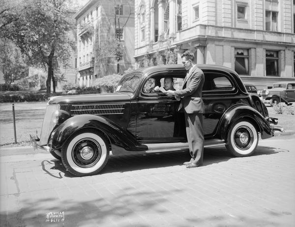 Salesman standing beside new Ford car, parked beside the U.W. YMCA, 740 Langdon Street, with Harry Stuhldreher, University of Wisconsin-Madison football coach, sitting inside.