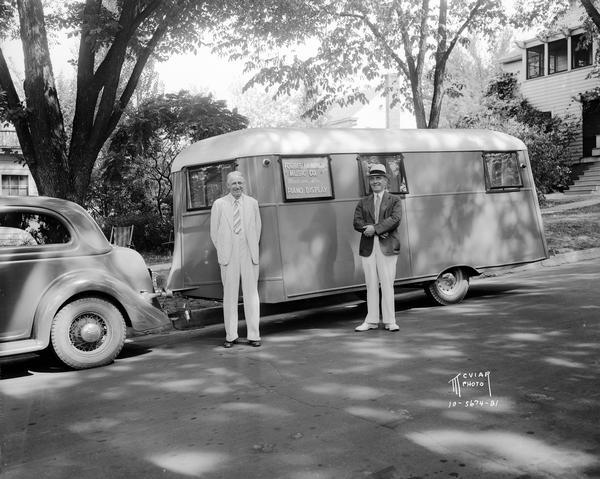 Two men standing in front of Forbes Meagher trailer with "Forbes Meagher Music Co., Madison, Wis. piano display" sign on side of trailer.