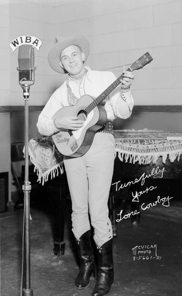 Man dressed as a cowboy playing a guitar and singing in front of WIBA microphone.