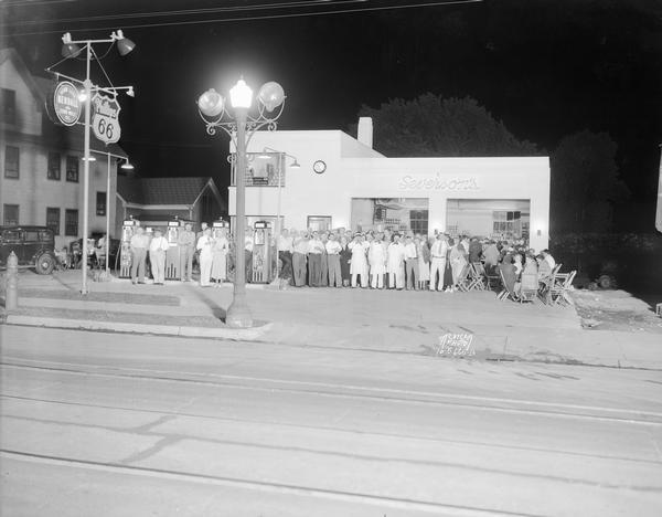 View across street of a large group of people posing in front of Severson's Phillips 66 Service Station, 2089 Atwood Avenue, during a summer evening beer party.