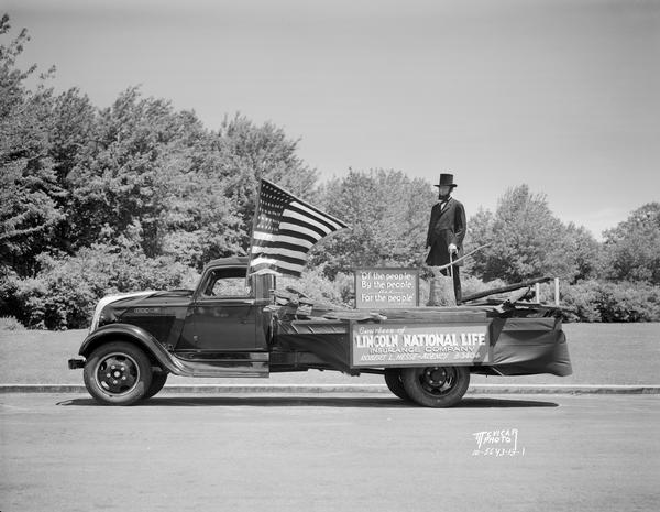 Abe Lincoln Float, G.A.R. parade. There is a man dressed as Abraham Lincoln standing in the back of a truck decorated with an American flag and sign saying: "Courtesy of Lincoln National Life Insurance Company."