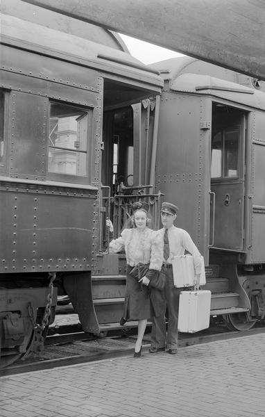 Manchesters Department Store model, wearing fur jacket hat and purse, posing with the conductor who is holding her luggage on the steps of a passenger train car.