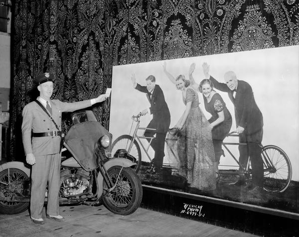 Roundy Coughlin in a police uniform, standing in front of a motorcycle and a life-sized poster of four movie stars on a tandem bicycle. This event occurred on the Orpheum Theatre stage, 216 State Street.