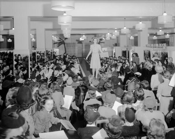 Women's style show with female model on the runway and women sitting in the audience at Manchester's Department Store.