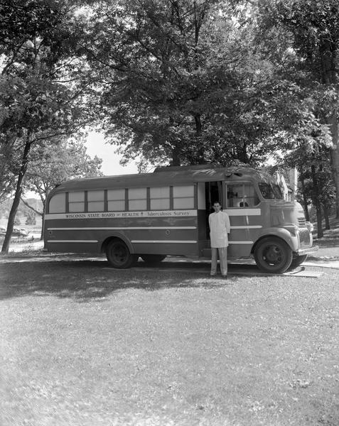 Bus containing mobile x-ray machine with sign, "Wisconsin State Board of Health, Tuberculosis Survey." A male technician is standing beside the bus. The bus is parked at the Oregon School for Girls.