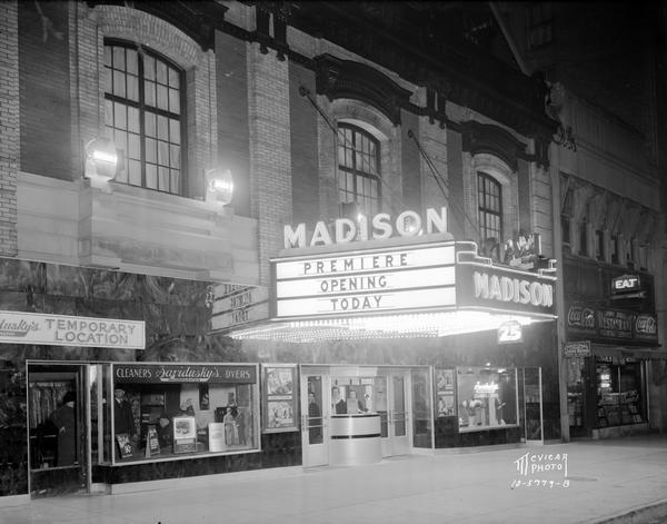 Night view of the Madison Theatre at 111 Monona Avenue. Two women are in the box office. The marquee reads "Premiere Opening Day." Jimmy Dodge's restaurant is to the right of the theater, and the temporary location of Savidusky's Cleaners and Dyers is on the left.