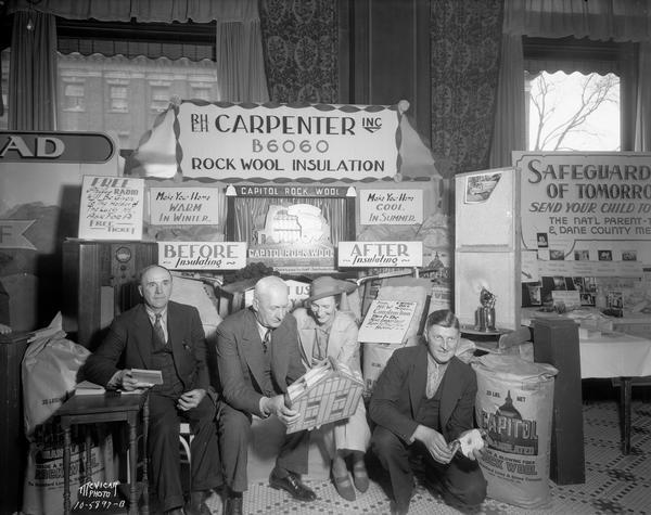 Three men and a woman looking at display materials in the R.H. and E.H. Carpenter, Inc. exhibit of Rock Wool insulation at the Home Show. Text on back of print reads: "Carpenter Real Estate, Insurance, Insulation Home Builders & Contractors, 102 W. Wilson St."