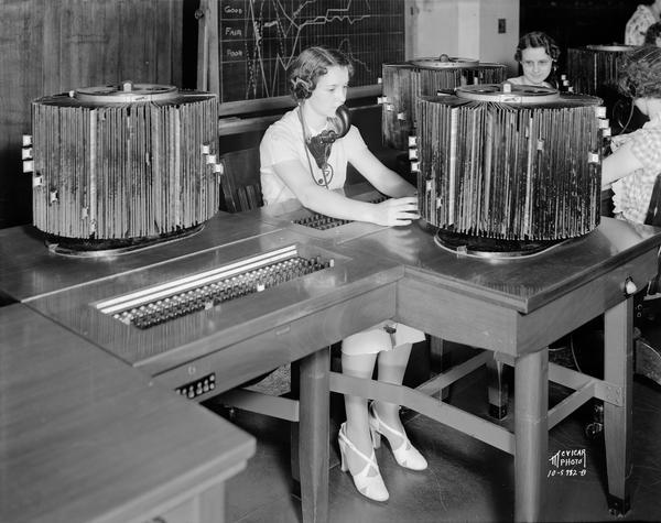 Women switchboard operators, wearing headsets with microphones at the Wisconsin Telephone Company, located at 17 S. Fairchild Street. There are rotary directory files on the tables.