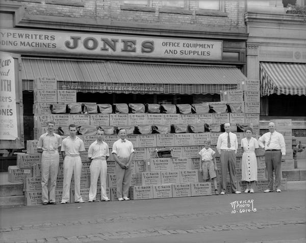 One child and seven adults standing in front of L.C. Smith typewriter boxes and covered typewriters stacked up in front of Jones Typewriter - Business Furniture Company located at 506 State Street.