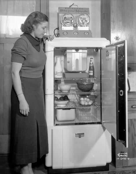 A woman is looking into a Westinghouse refrigerator full of food. There is an official test panel with 2 meters on top of the refrigerator.