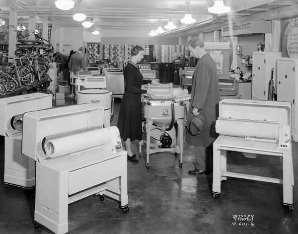Sales woman demonstrating a Maytag washing machine to a customer. In the foreground are Maytag Ironers (mangles) at Burdick & Murray, 26-30 N. Carroll Street. Bicycles are on display in the background on the left.