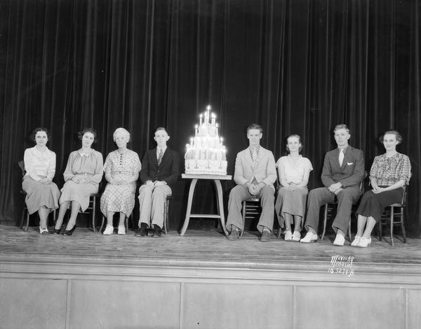 Seven East High School seniors and one woman sitting on stage with a tiered birthday cake with candles.