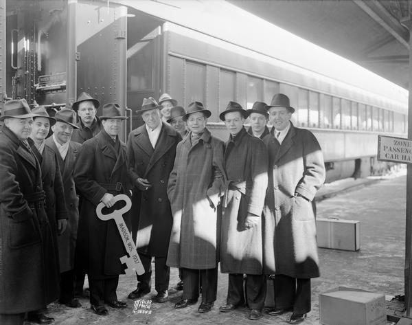 Rudy Vallee with eleven members of Sigma Alpha Epsilon fraternity wearing hats and coats and posing in front of Chicago and Northwestern railroad passenger car. Rudy Vallee is holding the key to the City of Madison.