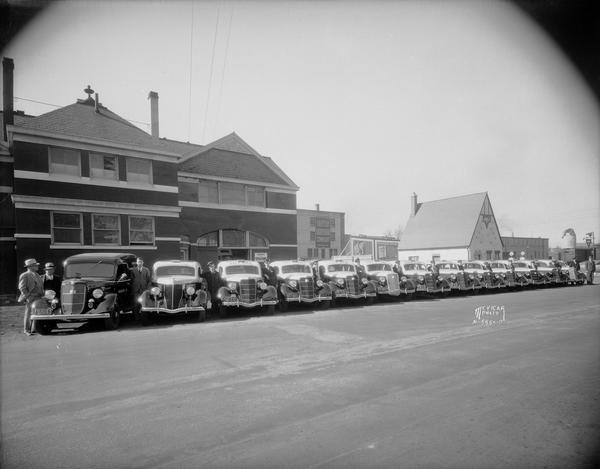 Lineup of 13 Ford City Car Company taxicabs and their drivers in front of the City Car Co., 601 - 603 West Washington Avenue. The view also contains Madison Hardware, 615 West Washington Avenue, and Fiore Service Station, 601 West Washington Avenue. The building was the Illinois Central Railroad Station.