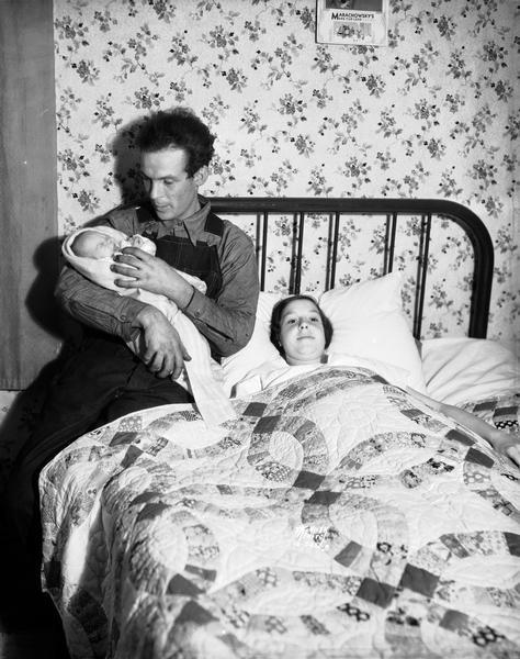 Arthur Parkins, Jr. holding surviving newborn twin baby, John Archie Parkins, following the death of twin baby James Burton Parkins. His wife Kathryn is lying in bed under a wedding ring quilt.