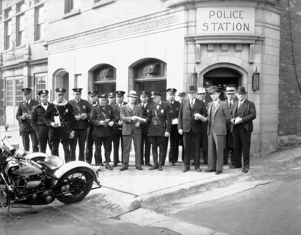 Group portrait of police officers receiving insurance policies in front of the Madison Police Station, 14-16 South Webster Street, with motorcycles parked at curb.