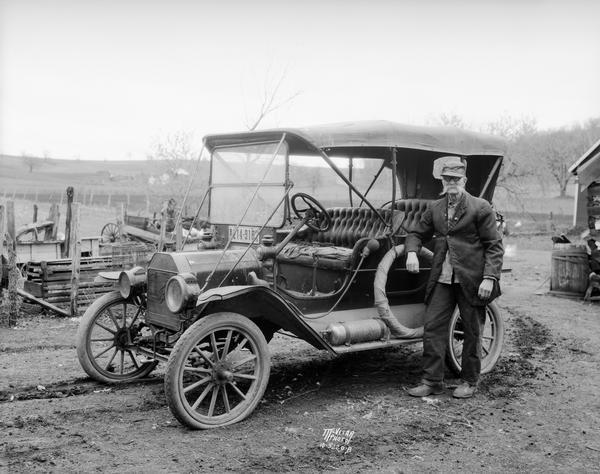 Man with bushy moustache standing next to a 1908 Ford Touring Car in a farm yard.