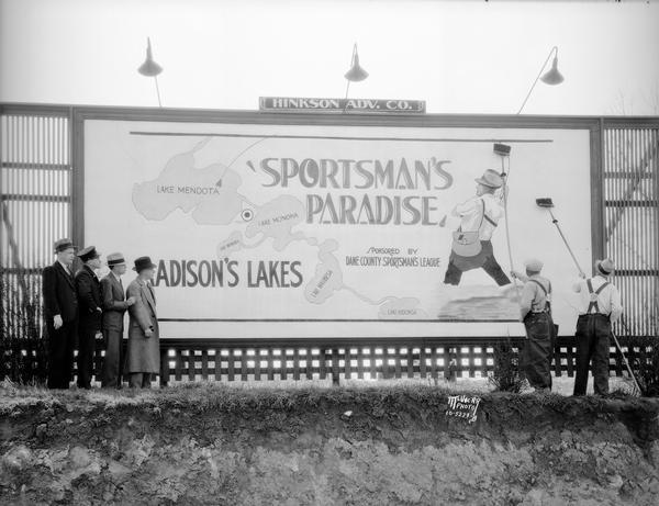 Four men are looking on during installation of a billboard designed by Hinkson Advertising Agency, at the corner of S. Park Street and Olin Avenue. The sign promotes Madison's lakes, with a depiction of a man fishing, and is titled "Sportsman's Paradise," sponsored by the Dane County Sportsman's League.