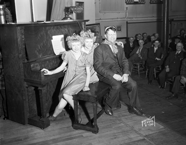The Gerke twins, Julie and Juliette, 11-years-old, are wearing matching dresses and bows in their hair. They are joined on the piano bench by a man wearing a bandana and business suit, during a Dane County Farm Bureau program at the Evangelical Lutheran Church. There is an audience in the background.