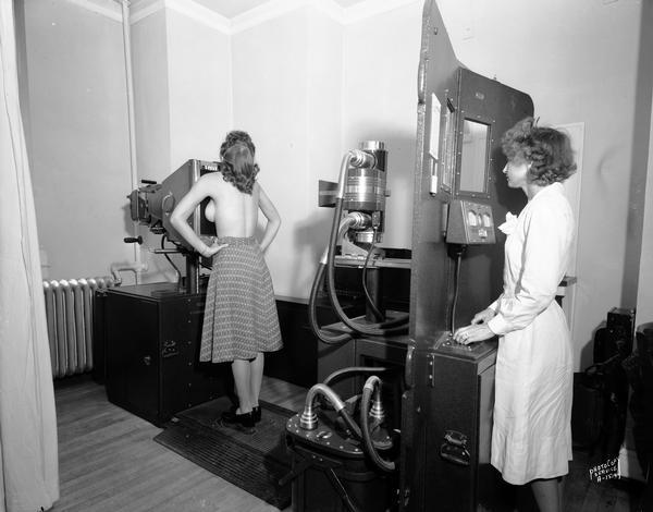 A woman, bare from the waist up, standing at the x-ray machine as she is receiving a chest x-ray taken by an attendant at the controls at the Wisconsin General Hospital.