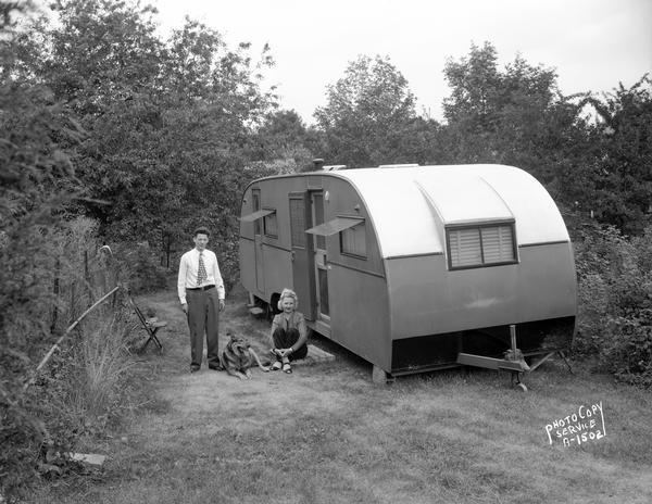 A man and woman posing with their dog outside their house trailer set in a secluded yard, surrounded by trees and bushes. Taken for Western Electric.