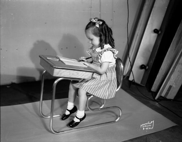 Pinafore-clad Carol Cochems, holding a pencil, with ringlets and a bow in her hair, reads in deep concentration at a desk-chair invented by H.M. Wood.
