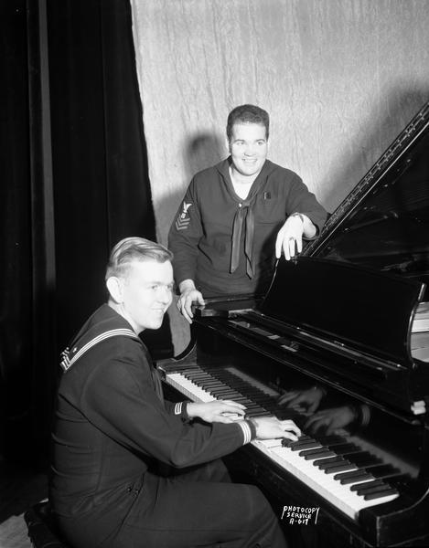 A U.S. Navy sailor playing piano while another sailor is leaning on the piano awaiting his time to sing.