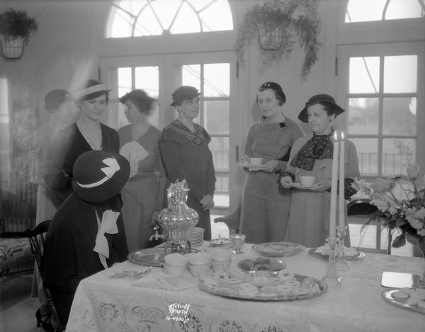 Women enjoying coffee, tea and pastries at the Maple Bluff Country Club. Left to right: Mrs. Oscar Tobaas, sitting at the table, with Mrs. H. Burnham Page standing behind her, Mrs. John St. John, Mrs. Mary Sayle Tegge, Mrs. Clifford Mathys, and Mrs. N.M. Schantz.