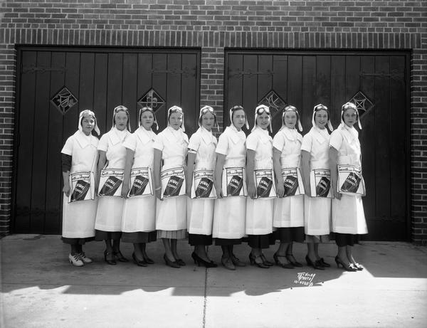 Ten hostesses dressed in white flying uniforms with helmets and goggles posing at the Kroger Grocery and Baking Co. air show and food exposition. They are from left: Louise Lambeck, Betty Withey, Jean Usher, Margaret Beihl, Betty McPeek, Ruth Ann Bailey, Mildred Withey, Louclaire Rapalje, Marion Grimm, and Helen Bonham.