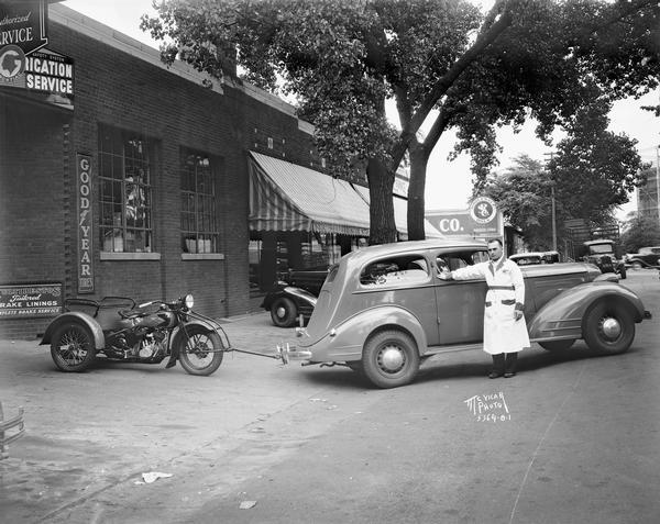 A man in a labcoat shows off a car hitched to a service motorcycle in front of Waters Motor's Co., 802 E. Washington Avenue.