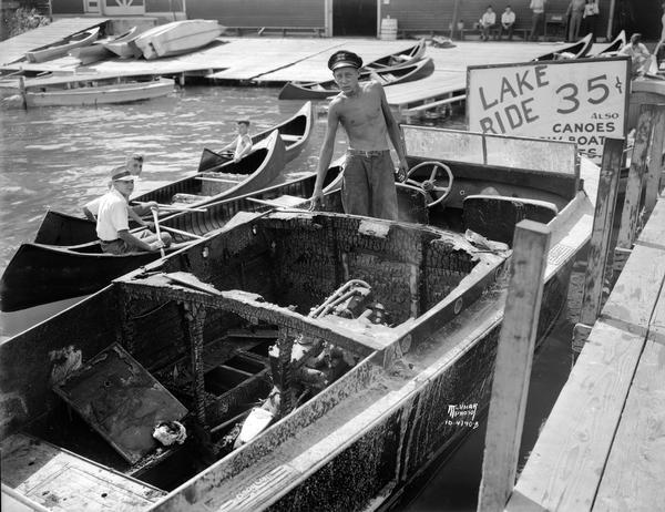 Canoers paddle by to observe Don Krueger standing in the bow of the burned-out "Baby Gar Wood" speedboat after six University of Wisconsin women students were forced to leap into Lake Mendota to escape the boat that had erupted into flames.