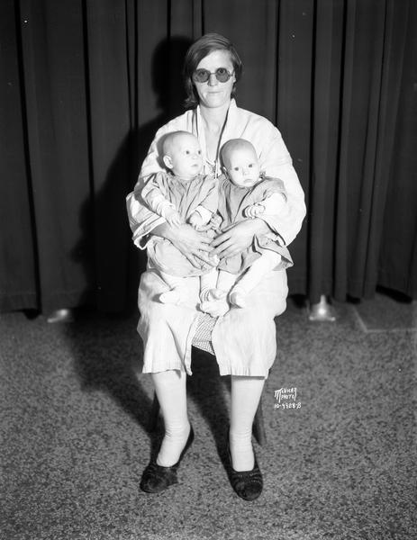 Portrait of Mrs. Morrow, 21-years-old, sitting and wearing dark eyeglasses due to a medical procedure for an eye infection. Morrow is from Mather, Wisconsin, and is holding her new twins, Geraldine Marie and Carolyn June, 4 months old. This is her third set of twins in three years.