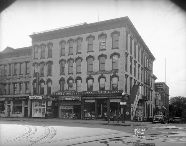 The Blied Building with storefronts, from left to right: Western Union, 21 W. Main Street, Madison Trust Company, 23 W. Main Street, Fanny Farmer, 25 W. Main Street, Band Box Millinery, 25 W. Main Street, Canton Restaurant, 25-27 W. Main Street, Forbes Meagher, 27 W. Main Street, and John J. Blied and Sons, 29 W. Main Street. Around the corner on South Carroll Street are Wisconsin Engraving Company, 109 S. Carroll Street, Press Club Tavern, 111 S. Carroll Street, and the Wisconsin State Journal, 115-123 S. Carroll Street.
