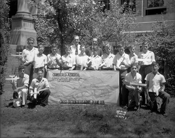 Group portrait of members of a vocational school model airplane builders club, each holding a model airplane with a diagram of a cambered aerofoil from a cross-section of a U.S. Army airplane rib.