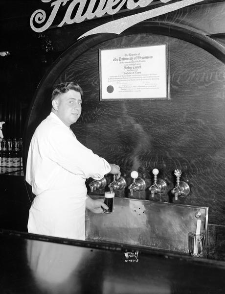 Apron-clad "Moon" Molinaro, Fauerbach's bartender, fills a stein of Fauerbach beer from the tap, with Arthur Towell's University of Wisconsin diploma on the side of the giant keg from which the beer is being drawn.