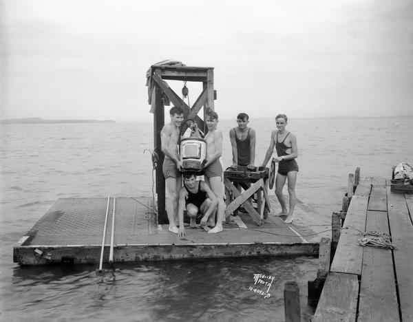 Five boys experimenting with diving apparatus on a diving raft. Left to right: Lyell Tullis, Edgar Tullis, and Jerry Randall fixing the diving head gear, with unidentified friends. Text on print reads: "Five boys with home-made diving bell on raft, Lake Mendota."