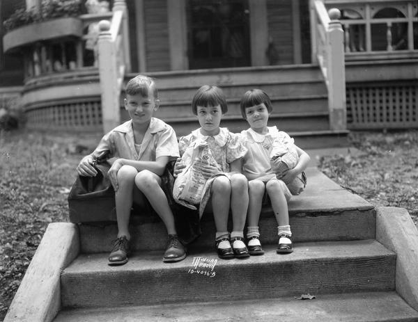 Two girls and a boy waiting on steps in front of their house to go to The Capital Times Kiddie Camp, clutching parcels that hold their camp clothes.